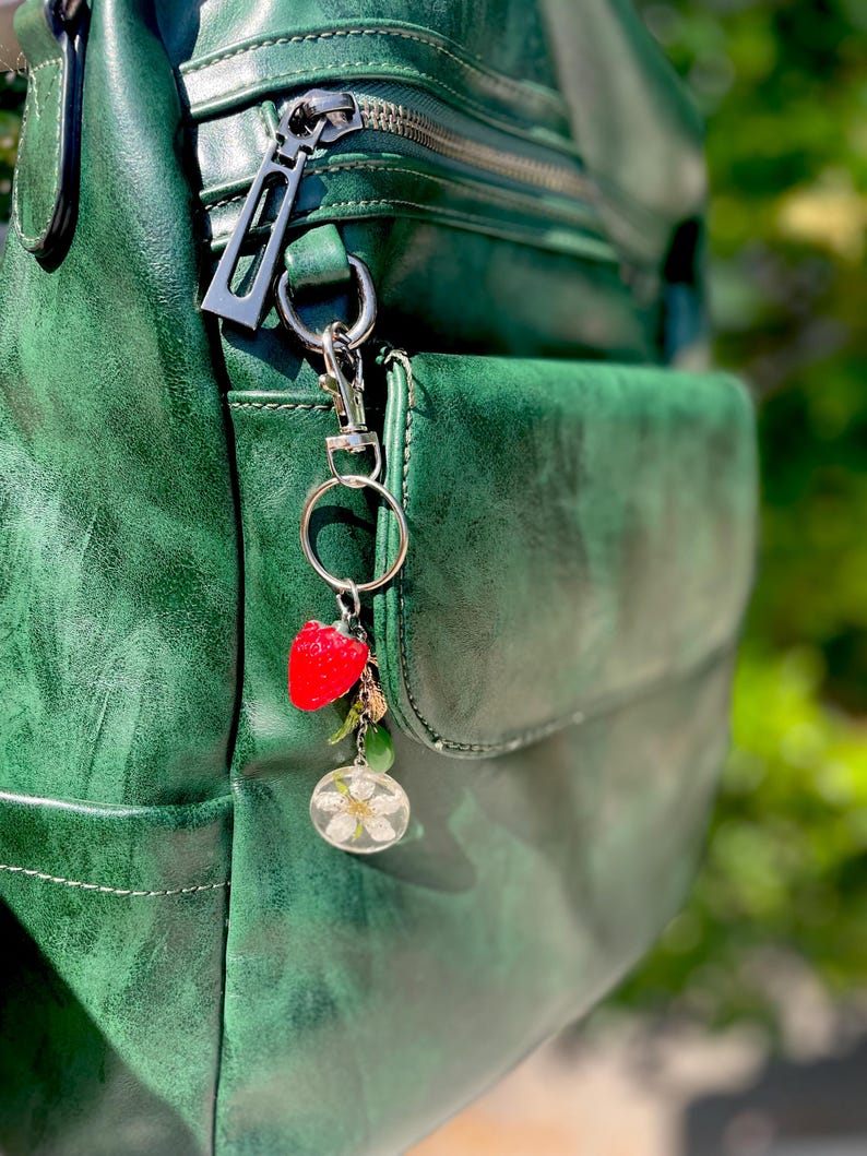 Peut inclure: Un sac &agrave; dos en cuir vert avec une fermeture &eacute;clair argent&eacute;e et un porte-cl&eacute;s contenant un charm en forme de fraise et un charm en r&eacute;sine transparente avec une fleur blanche. Le sac &agrave; dos a une finition textur&eacute;e.