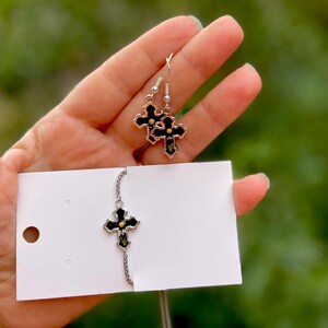 May include: A set of silver cross earrings and a silver cross necklace with black enamel and gold accents. The earrings and necklace feature a floral design.