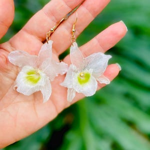 Pode incluir: Um par de brincos pendentes com berloques transl&uacute;cidos em forma de orqu&iacute;dea branca com centros verde-lim&atilde;o vibrantes. Os pingentes florais est&atilde;o presos a ganchos de metal dourado, criando uma apar&ecirc;ncia delicada e bot&acirc;nica para estes acess&oacute;rios.