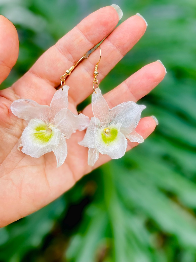 Pode incluir: Um par de brincos pendentes com berloques transl&uacute;cidos em forma de orqu&iacute;dea branca com centros verde-lim&atilde;o vibrantes. Os pingentes florais est&atilde;o presos a ganchos de metal dourado, criando uma apar&ecirc;ncia delicada e bot&acirc;nica para estes acess&oacute;rios.