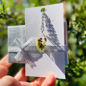 May include: A silver chain necklace with a clear teardrop pendant containing pressed flowers. The pendant is on a white card with a silver ribbon.