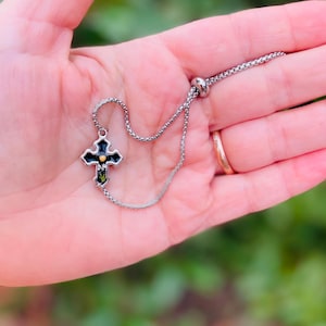 May include: A silver adjustable bracelet with a black cross charm featuring a gold center. The cross has a green and black floral design.