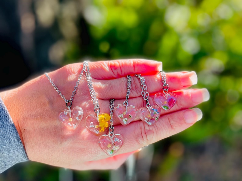 Puede incluir: Seis collares de plata con colgantes en forma de coraz&oacute;n transparentes llenos de flores prensadas. Los colgantes est&aacute;n llenos de flores de diferentes colores, incluyendo rosa, amarillo y blanco.