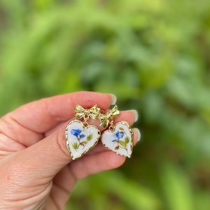 K&ouml;nnte beinhalten: Ein Paar goldene herzf&ouml;rmige Ohrringe mit wei&szlig;em Hintergrund und gepressten blauen und gr&uuml;nen Blumen. Jeder Ohrring hat oben eine goldene Schleife.