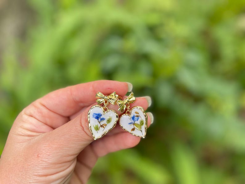 K&ouml;nnte beinhalten: Ein Paar goldene herzf&ouml;rmige Ohrringe mit wei&szlig;em Hintergrund und gepressten blauen und gr&uuml;nen Blumen. Jeder Ohrring hat oben eine goldene Schleife.