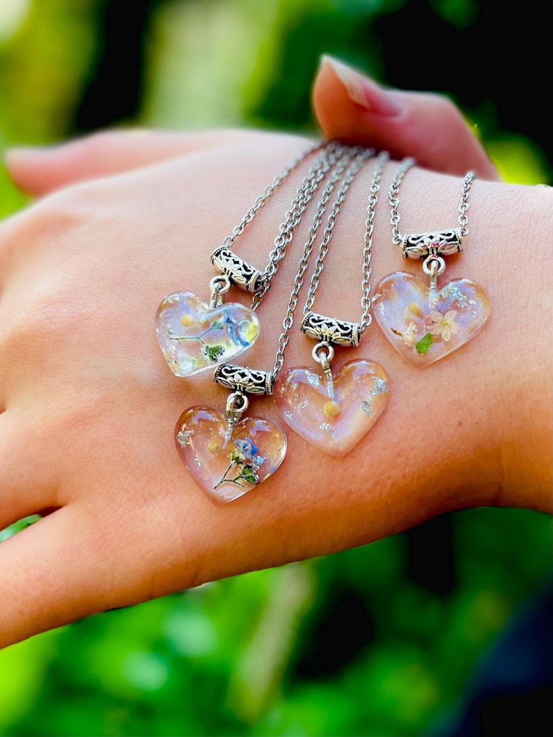 Puede incluir: Cuatro collares de plata con colgantes en forma de coraz&oacute;n transparentes llenos de flores prensadas. Las flores son blancas, amarillas y azules.