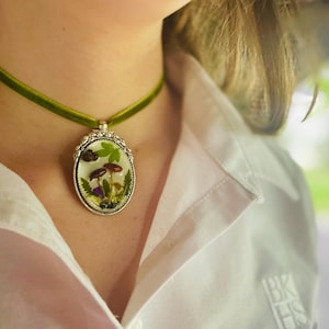 K&ouml;nnte beinhalten: Eine gr&uuml;ne Samt-Chokerkette mit einem silbernen ovalen Anh&auml;nger, der ein gepresstes Blumenmotiv aus Pilzen und Gr&uuml;n zeigt.