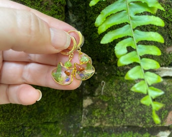 May include: Gold cat-shaped earrings with clear resin filled with green and purple dried flowers and small stones.