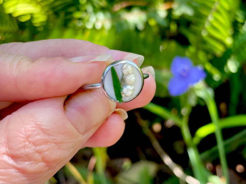 May include: A silver ring with a white and green resin inlay featuring a cluster of white lily of the valley flowers.