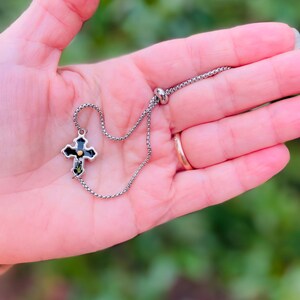 May include: A silver chain bracelet with a black cross charm featuring a gold center. The cross has a detailed design and is accented with small green stones.
