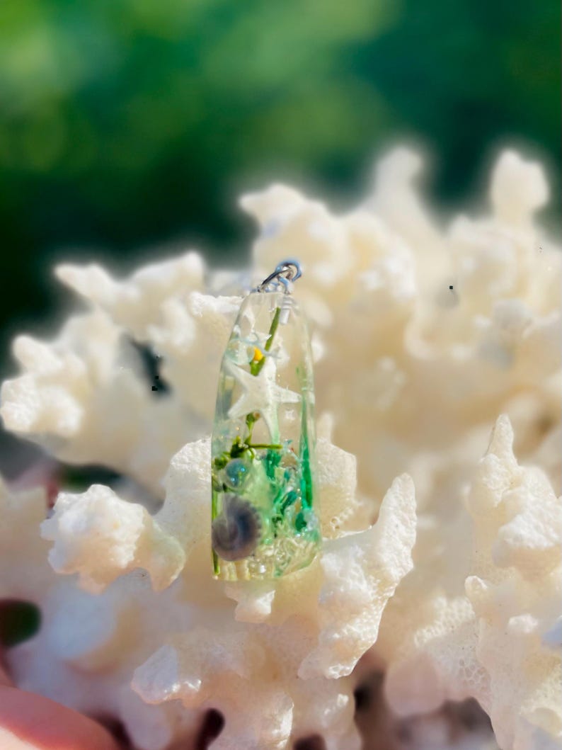 K&ouml;nnte beinhalten: Ein klarer Harzanh&auml;nger mit silberner &Ouml;se, der eine Miniatur-Unterwasserszene zeigt. Der Anh&auml;nger enth&auml;lt wei&szlig;e Seesterne, gr&uuml;ne Algen und eine kleine Muschel. Der Anh&auml;nger wird auf einem wei&szlig;en Korallenhintergrund pr&auml;sentiert.