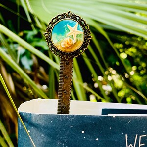 May include: A bronze metal bookmark with a decorative circular top featuring a beach scene with a starfish and seashells. The bookmark is inserted into a book with a dark blue cover. The bookmark has a ruler on the stem.