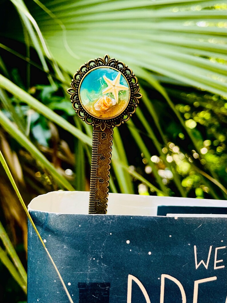 May include: A bronze metal bookmark with a decorative circular top featuring a beach scene with a starfish and seashells. The bookmark is inserted into a book with a dark blue cover. The bookmark has a ruler on the stem.