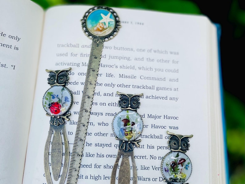 May include: A collection of decorative metal bookmarks with owl and beach-themed designs. The bookmarks feature intricate details, including a ruler, and are set against the backdrop of an open book.