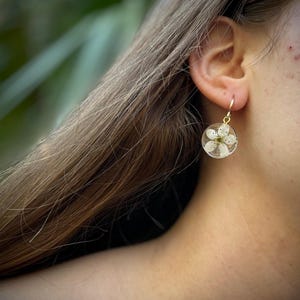 Puede incluir: Un par de pendientes de aro dorados con una flor blanca prensada dentro de un c&iacute;rculo de resina transparente.