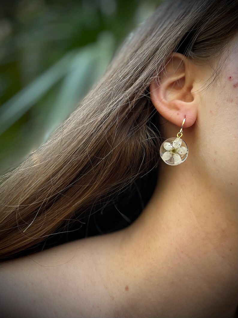 Puede incluir: Un par de pendientes de aro dorados con una flor blanca prensada dentro de un c&iacute;rculo de resina transparente.