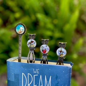 May include: A collection of decorative metal bookmarks and a ruler bookmark. The bookmarks feature owl and seashell designs with colorful accents. The bookmarks are placed on top of a blue book with the words "WE DREAM" on the cover.