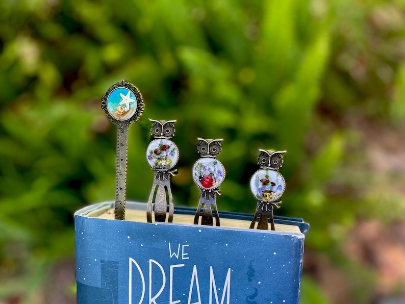 May include: A collection of decorative metal bookmarks and a ruler bookmark. The bookmarks feature owl and seashell designs with colorful accents. The bookmarks are placed on top of a blue book with the words "WE DREAM" on the cover.