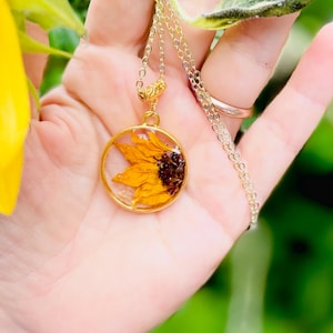 May include: A gold necklace with a circular pendant containing a preserved sunflower. The pendant showcases a yellow and brown sunflower, set in clear resin. The fine gold chain has a simple clasp, perfect for everyday wear.