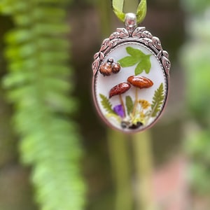 K&ouml;nnte beinhalten: Ein ovaler Silberanh&auml;nger mit wei&szlig;em Hintergrund, auf dem gepresste Blumen und Pilze zu sehen sind. Der Anh&auml;nger hat einen zarten silbernen Rahmen mit einem floralen Design.