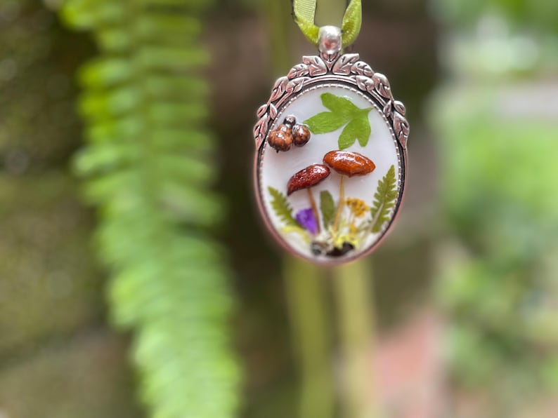 K&ouml;nnte beinhalten: Ein ovaler Silberanh&auml;nger mit wei&szlig;em Hintergrund, auf dem gepresste Blumen und Pilze zu sehen sind. Der Anh&auml;nger hat einen zarten silbernen Rahmen mit einem floralen Design.