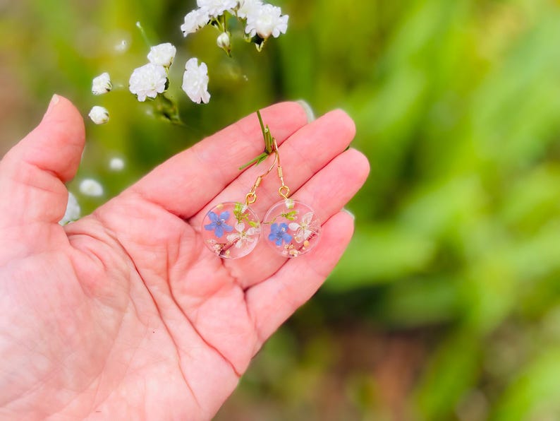 Puede incluir: Un par de pendientes de aro dorados con peque&ntilde;as flores azules y blancas prensadas en resina transparente.
