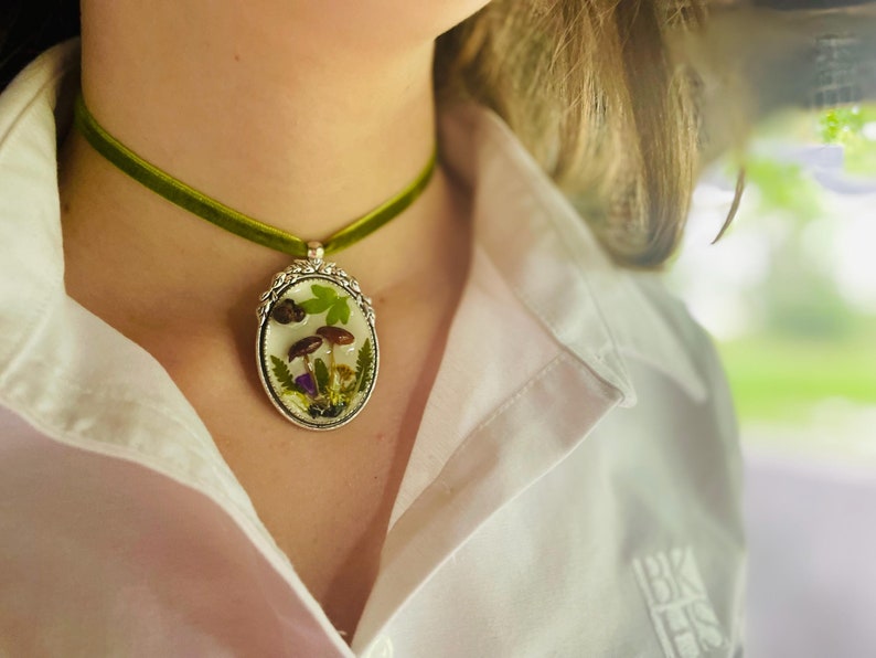 K&ouml;nnte beinhalten: Eine gr&uuml;ne Samt-Chokerkette mit einem silbernen ovalen Anh&auml;nger, der ein gepresstes Pilz- und Blumendesign zeigt.