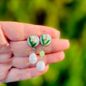 May include: A pair of handmade earrings featuring small, round, light blue resin settings with white flowers and green leaves. Each earring has a light green bead and a white pearl drop. The earrings are held in a hand.