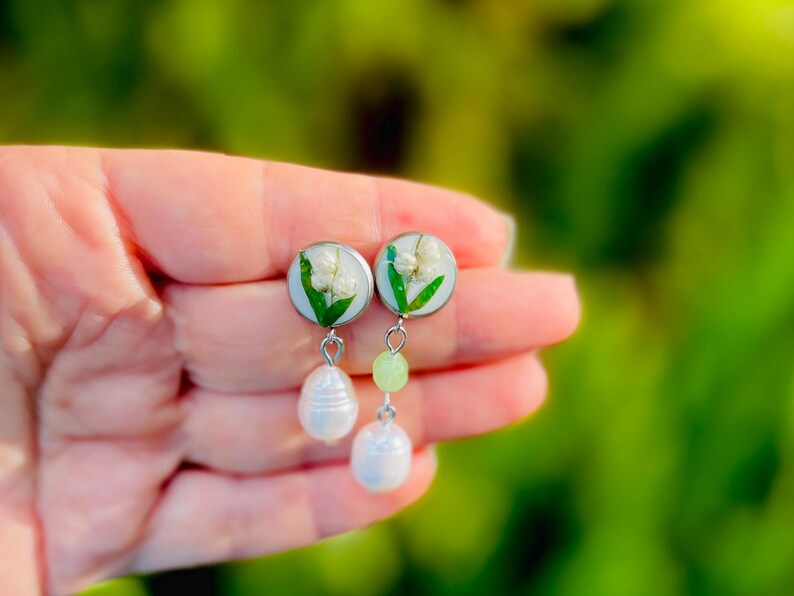 May include: A pair of handmade earrings featuring small, round, light blue resin settings with white flowers and green leaves. Each earring has a light green bead and a white pearl drop. The earrings are held in a hand.