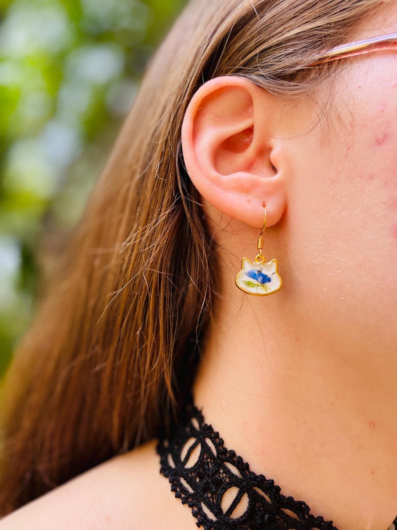 Puede incluir: Pendientes dorados con forma de gato con base de esmalte blanco y dise&ntilde;o floral azul. Los pendientes cuelgan de un gancho dorado. Los pendientes se llevan en la oreja de una persona.