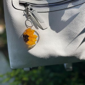 May include: A clear resin keyring with a bright yellow and brown sunflower design. The keyring is attached to a silver zip pull on a light grey leather bag. The bag has a textured surface and a silver zip.
