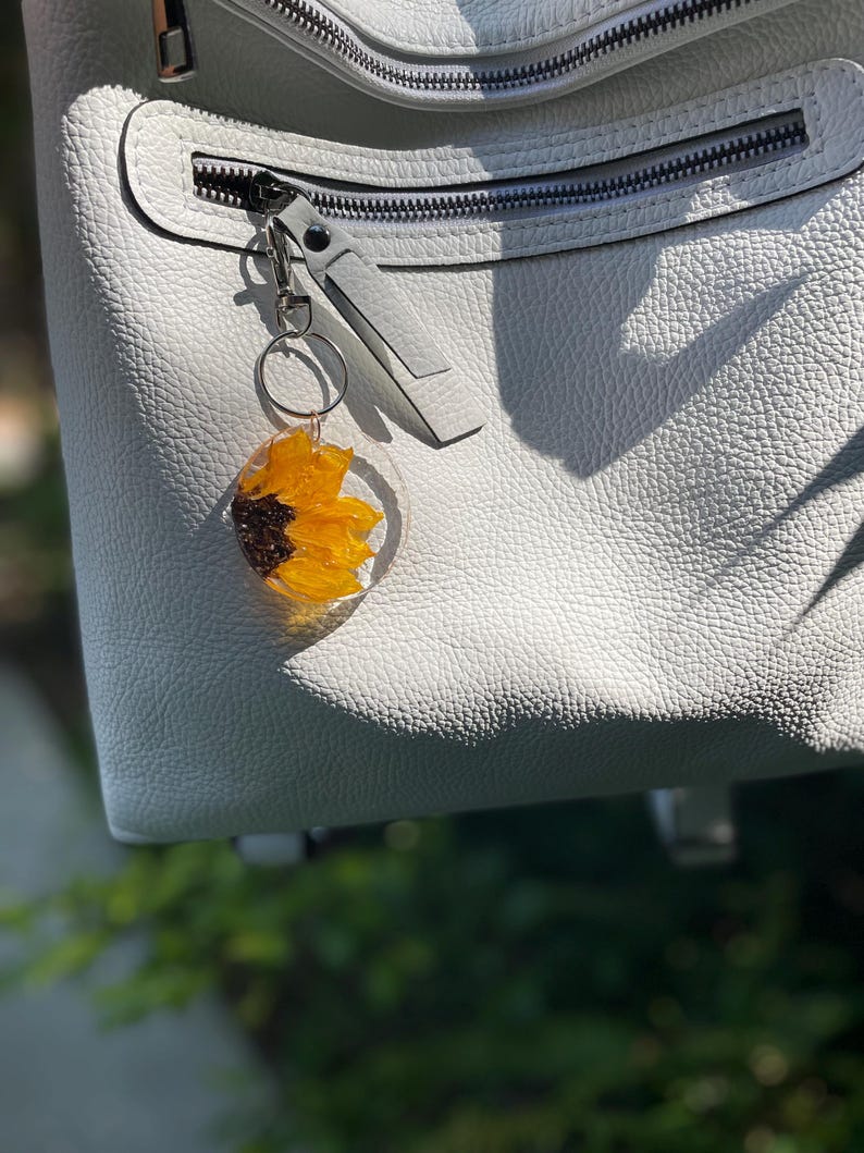 May include: A clear resin keyring with a bright yellow and brown sunflower design. The keyring is attached to a silver zip pull on a light grey leather bag. The bag has a textured surface and a silver zip.