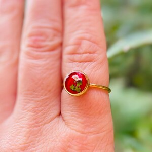 May include: A gold ring with a red resin setting featuring a small white flower and green leaves.