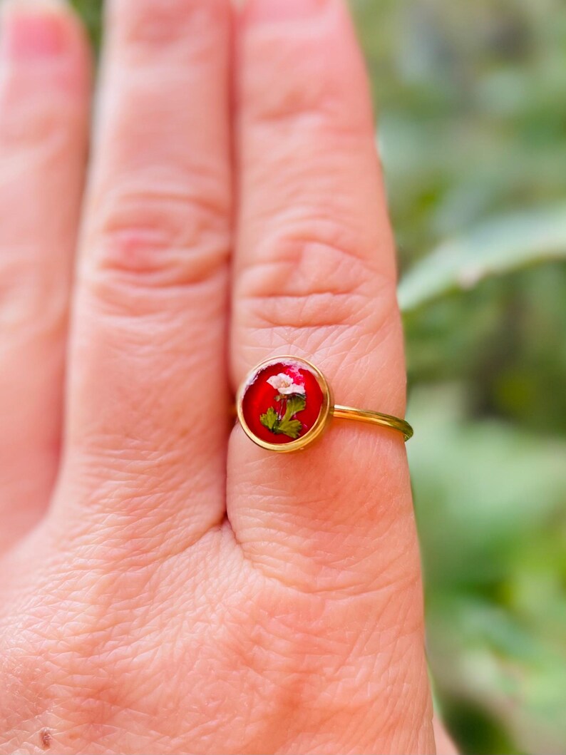 May include: A gold ring with a red resin setting featuring a small white flower and green leaves.