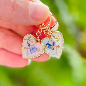 May include: A pair of gold heart-shaped earrings featuring a floral design. The earrings are made with real pressed flowers and have a delicate, feminine look.
