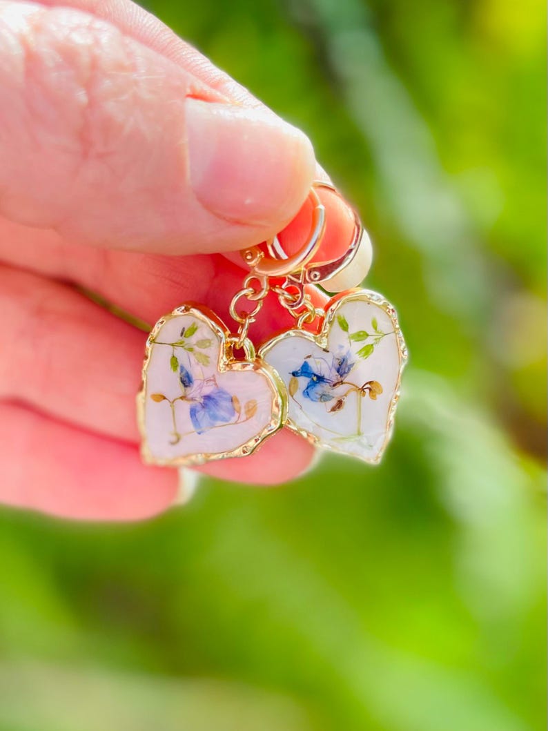 May include: A pair of gold heart-shaped earrings featuring a floral design. The earrings are made with real pressed flowers and have a delicate, feminine look.
