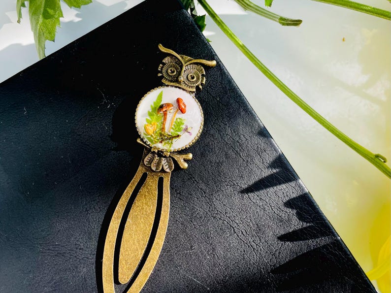 May include: A bronze owl bookmark with a round glass charm featuring a white background with a miniature mushroom scene. The scene includes brown and white mushrooms, green leaves, and a small white flower.