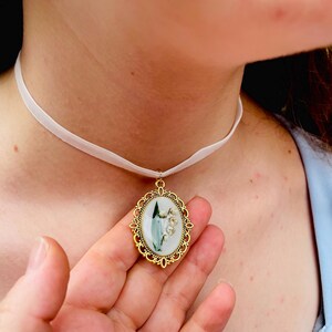 Pode incluir: Um colar de choker de cetim branco com um pingente oval dourado que apresenta uma flor de l&iacute;rio branca com folhas verdes.