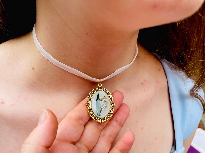 Pode incluir: Um colar de choker de cetim branco com um pingente oval dourado que apresenta uma flor de l&iacute;rio branca com folhas verdes.