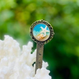 May include: A decorative bookmark featuring a beach scene with a white starfish, seashells, and blue water. The bookmark has a bronze-colored metal frame and a ruler on the bottom. The background is blurred green.