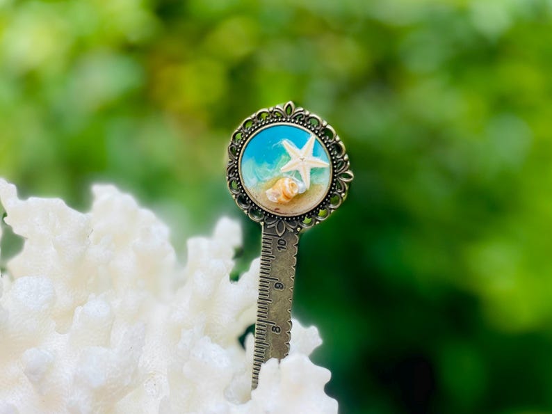 May include: A decorative bookmark featuring a beach scene with a white starfish, seashells, and blue water. The bookmark has a bronze-colored metal frame and a ruler on the bottom. The background is blurred green.