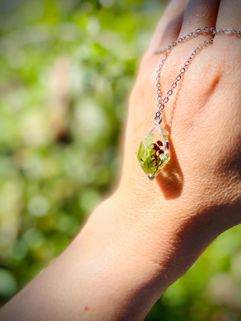 May include: A silver chain necklace with a clear pendant containing pressed green leaves and flowers.