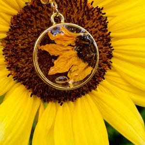 May include: A gold pendant necklace featuring a clear resin-filled circular charm with dried yellow sunflower petals. The pendant is set against a bright yellow sunflower with a dark brown centre.