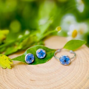 K&ouml;nnte beinhalten: Ein Set aus silbernem Schmuck mit einer blauen Blume, die in jedes St&uuml;ck gepresst ist. Das Set enth&auml;lt einen Ring und zwei Ohrringe.