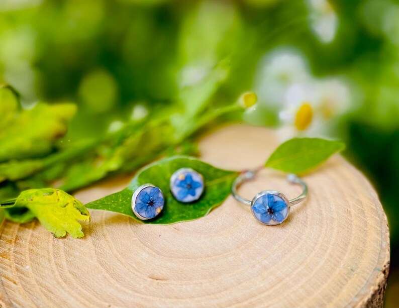K&ouml;nnte beinhalten: Ein Set aus silbernem Schmuck mit einer blauen Blume, die in jedes St&uuml;ck gepresst ist. Das Set enth&auml;lt einen Ring und zwei Ohrringe.