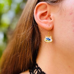 Puede incluir: Pendientes dorados con forma de gato con base de esmalte blanco y dise&ntilde;o floral azul. Los pendientes cuelgan de un gancho dorado. Los pendientes los lleva una persona con el pelo largo y casta&ntilde;o.
