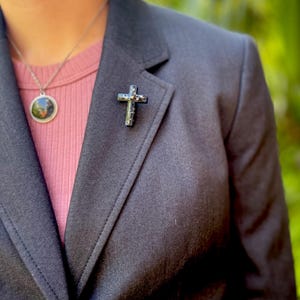 May include: A black blazer with a silver cross pin on the lapel. The blazer is worn over a pink shirt.