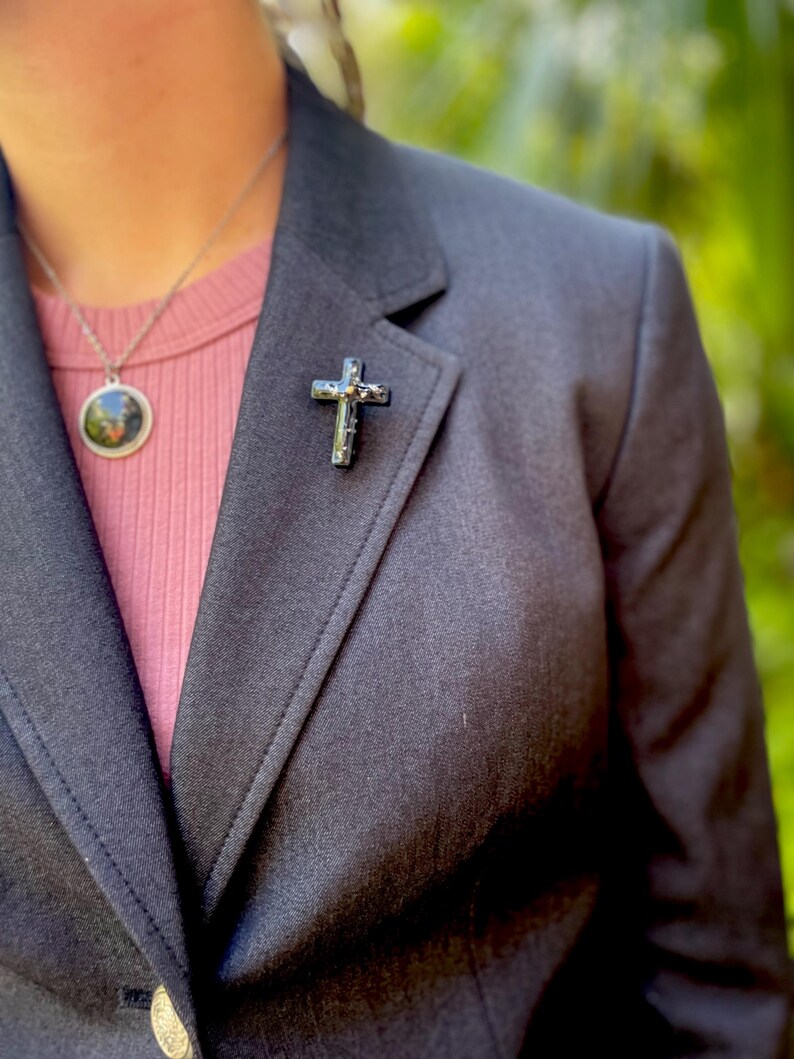 May include: A black blazer with a silver cross pin on the lapel. The blazer is worn over a pink shirt.