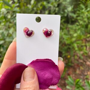 May include: A pair of heart-shaped earrings with pink and gold accents. The earrings are made of clear resin and contain dried rose petals.