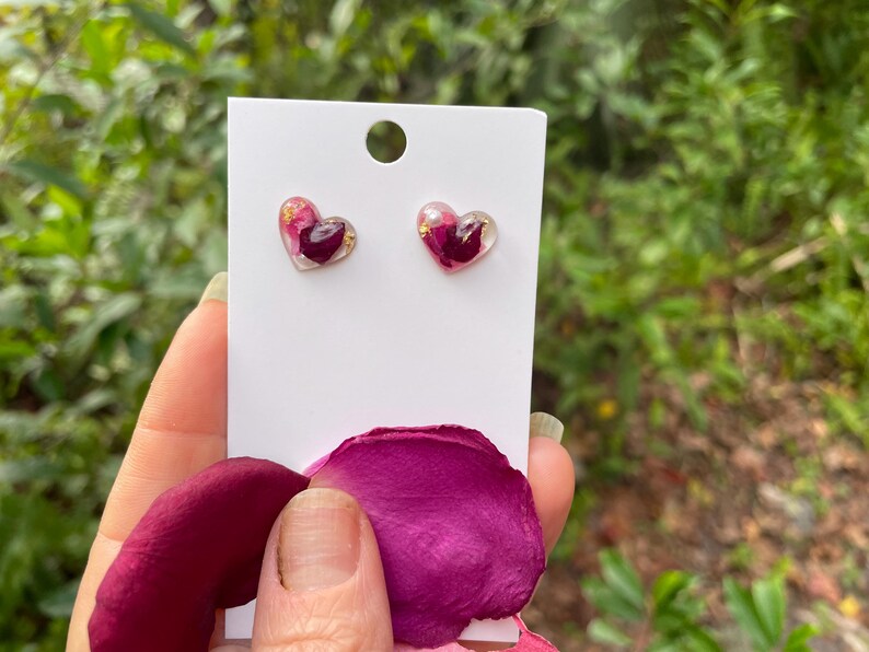 May include: A pair of heart-shaped earrings with pink and gold accents. The earrings are made of clear resin and contain dried rose petals.